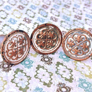 May include: Three antique-style cabinet knobs with a distressed finish. The knobs have a circular design with an ornate, symmetrical pattern in shades of white, copper, and gray. The knobs are made of metal and have a rustic, vintage aesthetic.