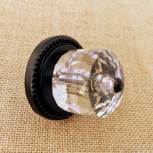 May include: A clear, faceted crystal cabinet knob with a black, textured base. The knob has a round shape and is designed for furniture. The crystal reflects light, creating a sparkling effect. The base has a decorative, ridged edge.