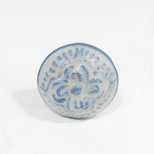 May include: A white ceramic knob with a blue floral design. The knob is round and has a raised design.