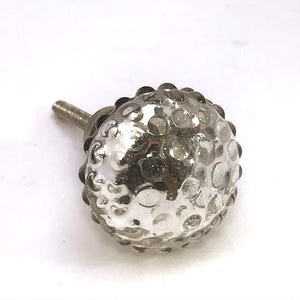 May include: A silver-colored, spherical cabinet knob with a textured surface and a threaded post. The knob has a dimpled pattern, and the post is made of metal. The knob is designed for use on drawers or cabinets.