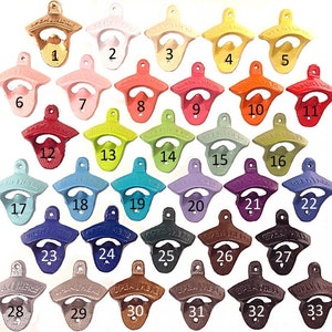 May include: A collection of cast iron bottle openers in various colors, including gold, white, pink, red, green, blue, and brown. Each opener is numbered and features the text "OPEN HERE".
