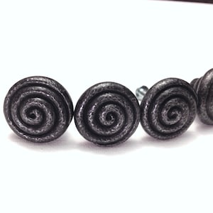 May include: Four dark gray metal cabinet knobs with a spiral design. The knobs are round and have a textured surface. The knobs are arranged in a row against a white background.