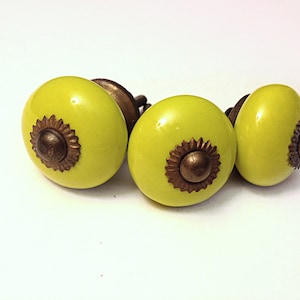 May include: Four lime green ceramic cabinet knobs with bronze-colored metal accents. Each knob has a rounded shape with a decorative bronze floral design in the center. The knobs are arranged on a white surface.