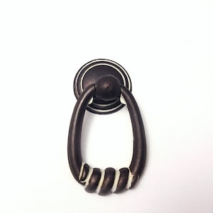 May include: A dark bronze-colored cabinet pull with an oval handle and a circular backplate. The handle features a decorative, twisted design with white accents. This hardware is suitable for drawers and cabinets.