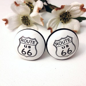 May include: Two white ceramic cabinet knobs with black trim. Each knob features a black and white Route 66 sign with the text "ROUTE US 66".