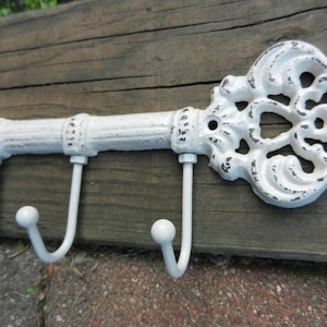 May include: A white, key-shaped wall hook with three curved hooks. The decorative key has an ornate design and a distressed finish, mounted on a wooden plank. The key is approximately 12 inches long.