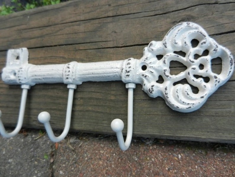 May include: A white, key-shaped wall hook with three curved hooks. The decorative key has an ornate design and a distressed finish, mounted on a wooden plank. The key is approximately 12 inches long.