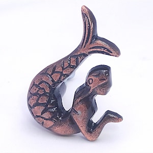 May include: Bronze-colored mermaid figurine with a detailed tail and a seated pose. The mermaid's tail is arched, and the figure has a textured, antique finish. The sculpture is a decorative item.