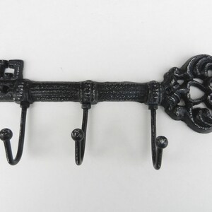 May include: Black cast iron key-shaped wall hook with three curved hooks. The key's head features a decorative, ornate design. The key's shaft has a textured, ridged appearance. The hooks have a rounded ball at the end.