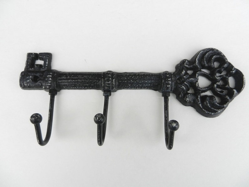May include: Black cast iron key-shaped wall hook with three curved hooks. The key's head features a decorative, ornate design. The key's shaft has a textured, ridged appearance. The hooks have a rounded ball at the end.