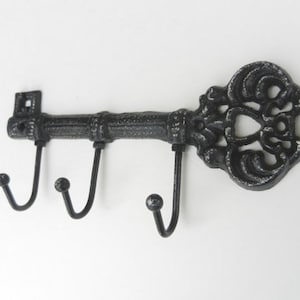 May include: A decorative, cast iron key-shaped wall hook with three curved hooks. The key is black with a distressed finish, featuring ornate detailing on the head. The hooks are evenly spaced along the key's shaft.
