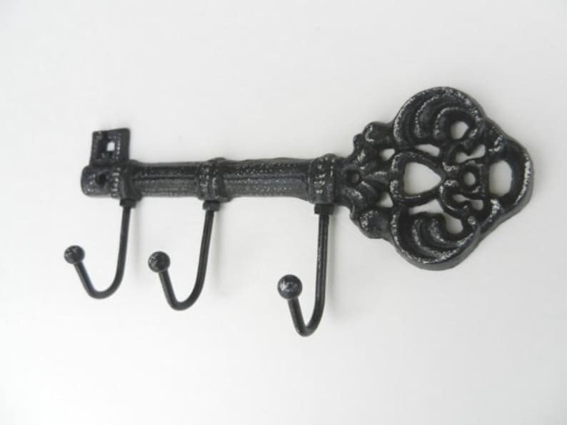 May include: A decorative, cast iron key-shaped wall hook with three curved hooks. The key is black with a distressed finish, featuring ornate detailing on the head. The hooks are evenly spaced along the key's shaft.