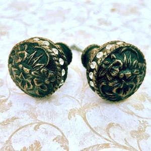 May include: Two antique-style cabinet knobs. The knobs are dark green with gold accents and a floral design. Each knob is adorned with small, clear crystals around the edge. These decorative knobs are perfect for adding a vintage touch to furniture.