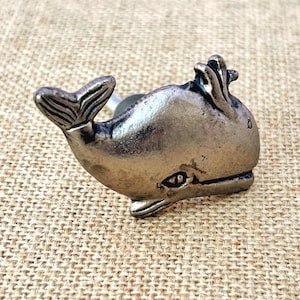 May include: A silver-toned whale-shaped cabinet knob. The knob features a detailed tail and flipper design. The whale is facing to the right. The knob is made of metal and has a slightly distressed finish. The knob is designed for use on drawers or cabinets.