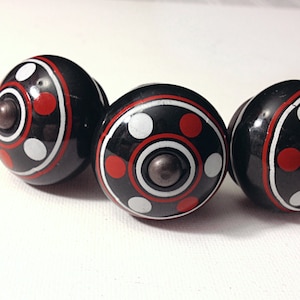 May include: Three black ceramic knobs with a white and red geometric design. The knobs have a circular shape and a raised center.
