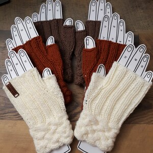May include: A collection of fingerless gloves in various colors, including cream, rust, and brown. The gloves are displayed on cardboard hand cutouts, showcasing their knitted texture and cable knit detailing. A small leather tag is attached to one of the cream gloves.