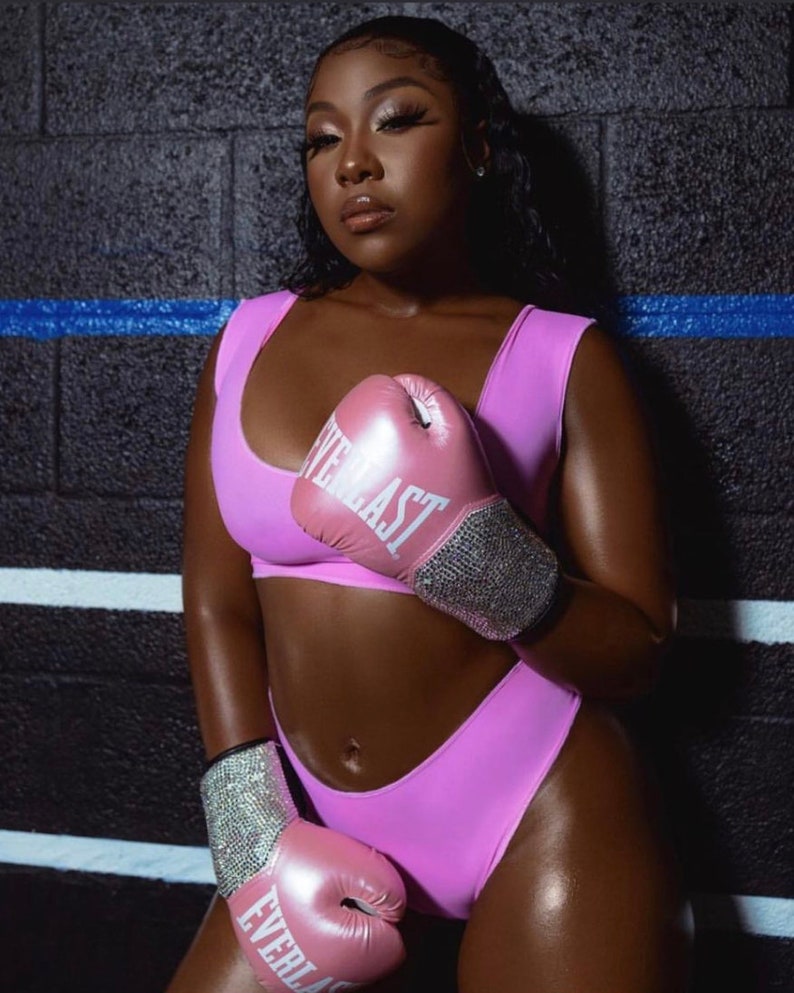 May include: A woman wearing a pink sports bra and matching shorts, holding pink boxing gloves with rhinestones. The gloves have the word "EVERLAST" printed on them.