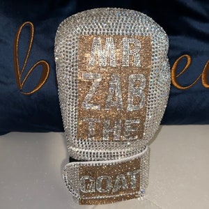 May include: A white boxing glove covered in clear and gold rhinestones. The glove has a rectangular patch on the front with the text "MR ZAB THE GOAT" in gold rhinestones.