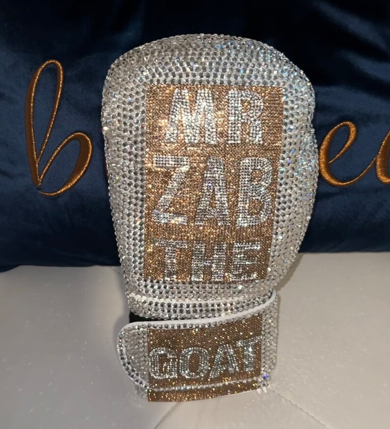 May include: A white boxing glove covered in clear and gold rhinestones. The glove has a rectangular patch on the front with the text "MR ZAB THE GOAT" in gold rhinestones.