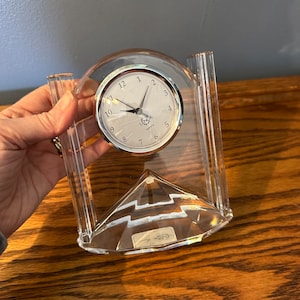 May include: A clear crystal clock with a silver face and black hands. The clock is on a clear crystal base with a pyramid shape. The clock face says "Levox Quartz".