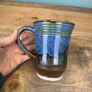 May include: A handmade ceramic mug with a blue and brown glaze. The mug has a rounded handle and a white base. The blue glaze has vertical streaks, and the rim has horizontal lines. The mug is approximately 4 inches tall.