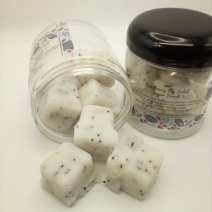 May include: A glass jar filled with white sugar scrub cubes with red seeds. The jar is labeled "Cranberry Fig Solid Sugar Scrub". The ingredients are listed on the label. The scrub is made with coconut oil, palm oil, sugar, and other natural ingredients.
