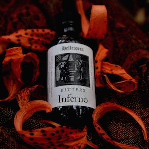 May include: A dark glass bottle of "Inferno" bitters with a white label featuring a black and white illustration. The bottle is surrounded by orange and brown flower petals, creating a moody, autumnal aesthetic.