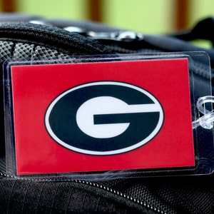May include: A red luggage tag with a black and white 'G' inside a black oval. The tag is attached to a black backpack.