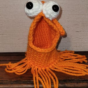 May include: A handmade orange crochet monster with large white eyes and black pupils. The monster has an open mouth and fringe around the base. The creature is sitting on a wooden surface.