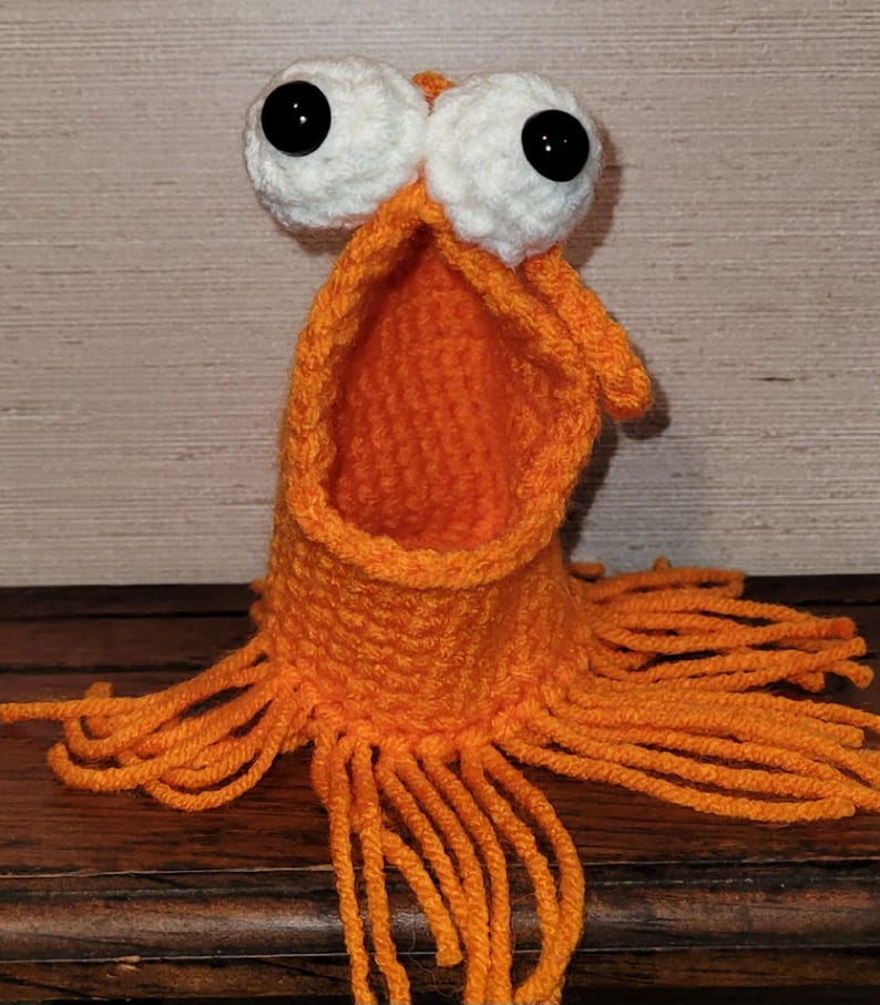 May include: A handmade orange crochet monster with large white eyes and black pupils. The monster has an open mouth and fringe around the base. The creature is sitting on a wooden surface.