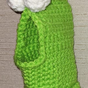 May include: A handmade, bright green crochet frog-like creature with white eyes and black pupils. The whimsical design includes a fringed bottom and a loop for hanging. The item is likely a decorative piece or a toy.