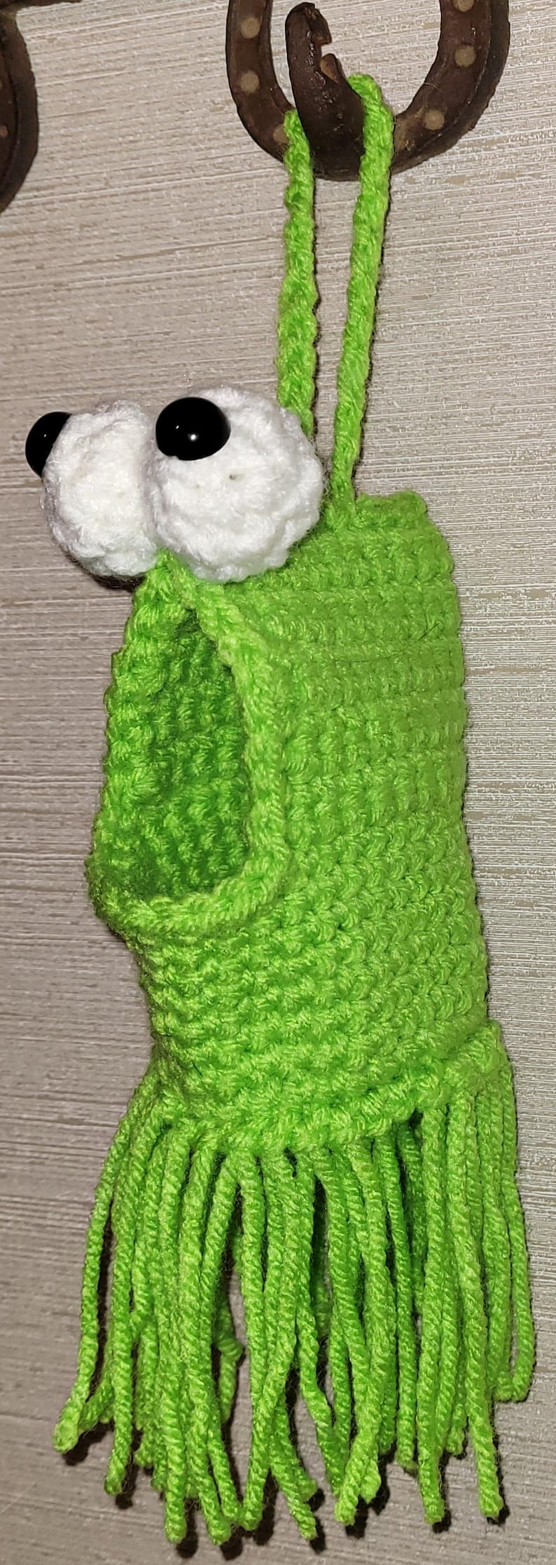 May include: A handmade, bright green crochet frog-like creature with white eyes and black pupils. The whimsical design includes a fringed bottom and a loop for hanging. The item is likely a decorative piece or a toy.