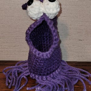 May include: A handmade purple crochet monster with large white eyes and black pupils. The monster has an open mouth and fringe at the bottom. The item is made of yarn and is sitting on a wooden surface.