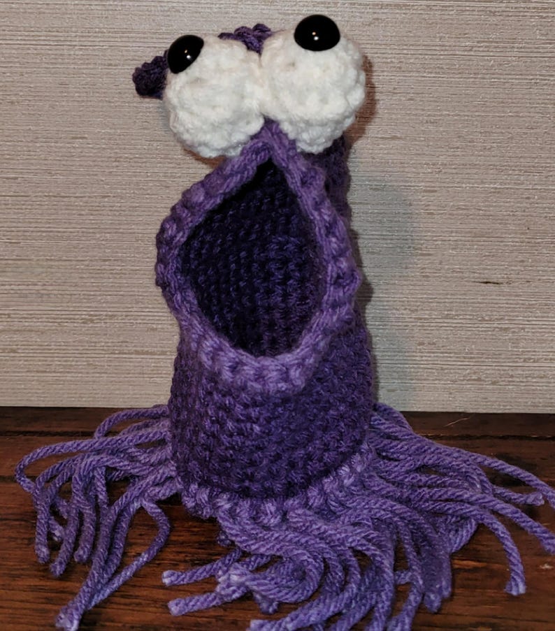 May include: A handmade purple crochet monster with large white eyes and black pupils. The monster has an open mouth and fringe at the bottom. The item is made of yarn and is sitting on a wooden surface.