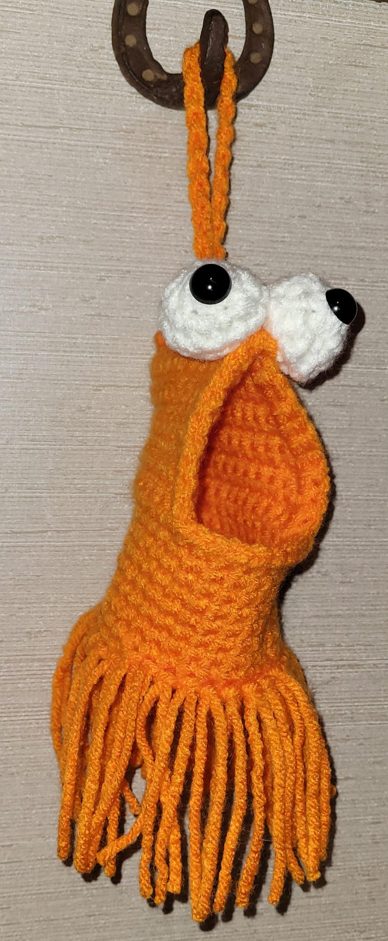 May include: A handmade orange crochet monster with large white eyes and black pupils. The monster has a wide open mouth and fringe at the bottom. It is hanging from a horseshoe.
