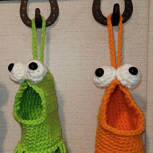May include: Two crocheted hanging pouches, one lime green and one orange, each featuring large white eyes with black pupils and a wide open mouth. The pouches have a fringed bottom edge and are suspended by matching yarn loops from a metal rack.