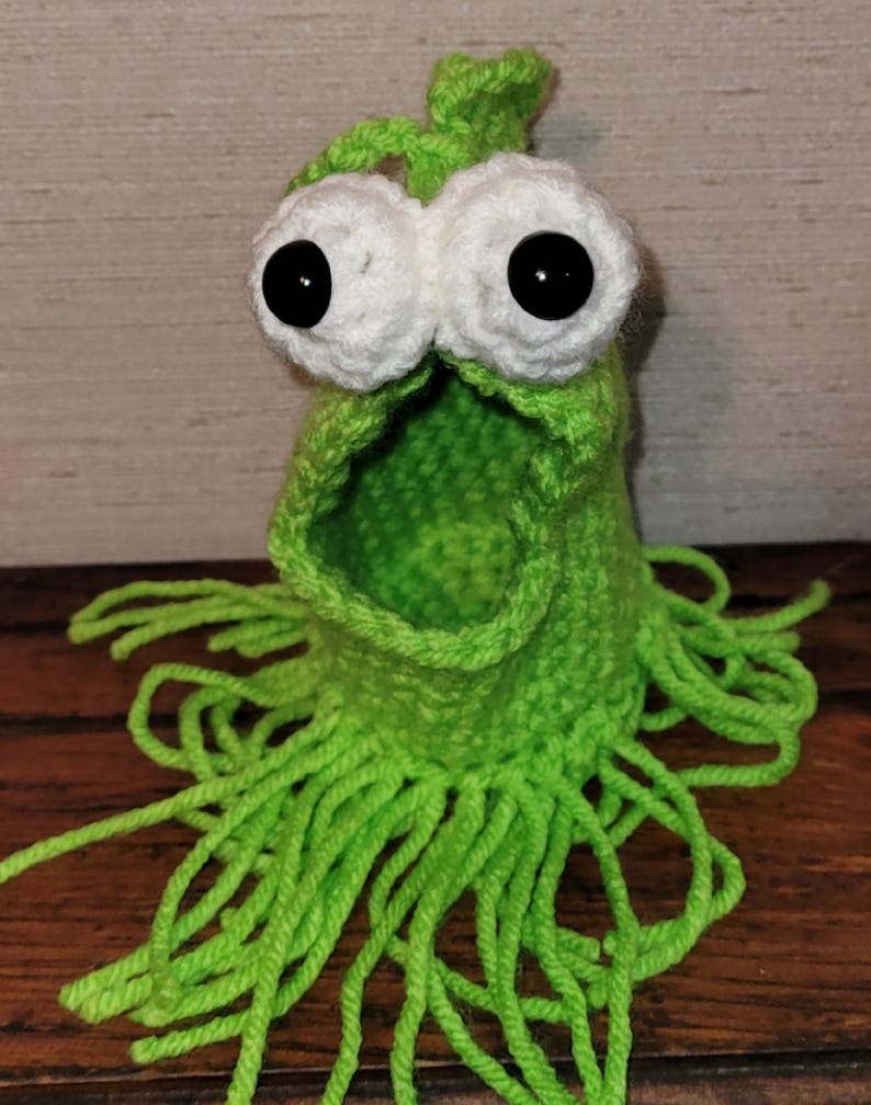 May include: A handmade, green crocheted monster with large white eyes and black pupils. The monster has an open mouth and green fringe around the bottom. The item is made of yarn and is likely a toy or decorative item.