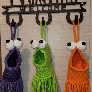 May include: Three handmade crochet monster-like wall pockets in purple, green, and orange. Each has large googly eyes and a fringe bottom. They hang from a metal welcome sign with horseshoe hooks.