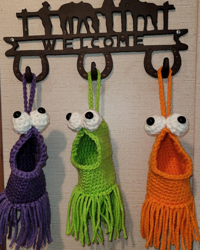 May include: Three handmade crochet monster-like wall pockets in purple, green, and orange. Each has large googly eyes and a fringe bottom. They hang from a metal welcome sign with horseshoe hooks.