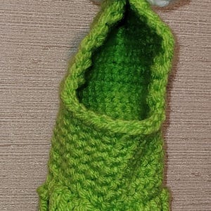 May include: A quirky, handmade green crochet monster with white eyes and black pupils. The monster has a fringed bottom and is suspended from a horseshoe. This unique item is perfect for Halloween or as a fun decorative piece.