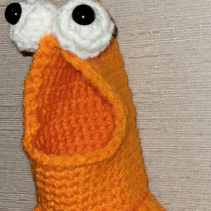 May include: A quirky, handmade orange crochet creature with large white-rimmed eyes and black pupils. The creature has a fringed bottom and is suspended from a horseshoe. The craftwork is detailed and the colour is vibrant.