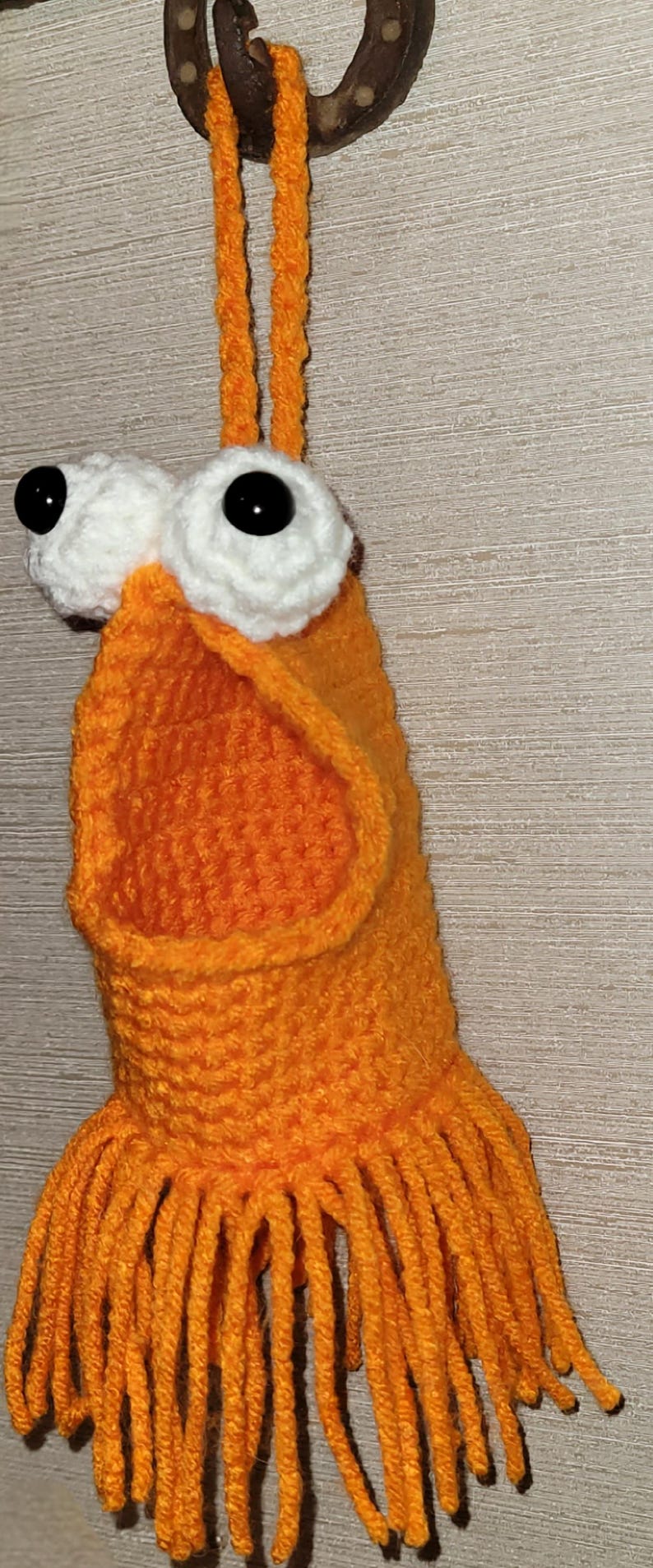 May include: A quirky, handmade orange crochet creature with large white-rimmed eyes and black pupils. The creature has a fringed bottom and is suspended from a horseshoe. The craftwork is detailed and the colour is vibrant.