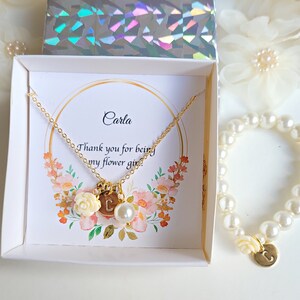 K&ouml;nnte beinhalten: Eine wei&szlig;e Box mit einer goldenen Halskette und einem Perlenarmband. Die Halskette hat einen Anh&auml;nger mit dem Buchstaben "C" und einer kleinen wei&szlig;en Blume. Das Armband hat ebenfalls einen Anh&auml;nger mit dem Buchstaben "C". Die Box enth&auml;lt eine Karte mit der Aufschrift "Carla, Danke, dass du meine Blumenm&auml;dchen bist".