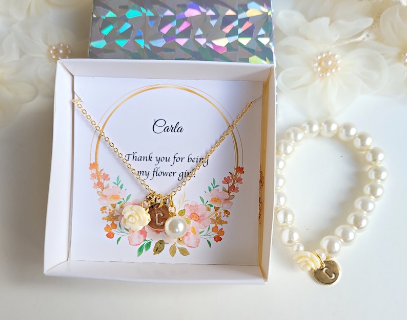 K&ouml;nnte beinhalten: Eine wei&szlig;e Box mit einer goldenen Halskette und einem Perlenarmband. Die Halskette hat einen Anh&auml;nger mit dem Buchstaben "C" und einer kleinen wei&szlig;en Blume. Das Armband hat ebenfalls einen Anh&auml;nger mit dem Buchstaben "C". Die Box enth&auml;lt eine Karte mit der Aufschrift "Carla, Danke, dass du meine Blumenm&auml;dchen bist".