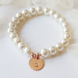 May include: A white pearl bracelet with a rose gold charm featuring the letter "S".