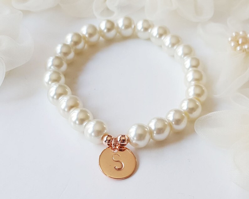 May include: A white pearl bracelet with a rose gold charm featuring the letter "S".