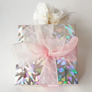 K&ouml;nnte beinhalten: Eine Geschenkbox, verpackt in schimmerndes silbernes Papier mit einer rosa Schleife aus Satin. Die Box ist mit einem wei&szlig;en Blumenarrangement verziert.