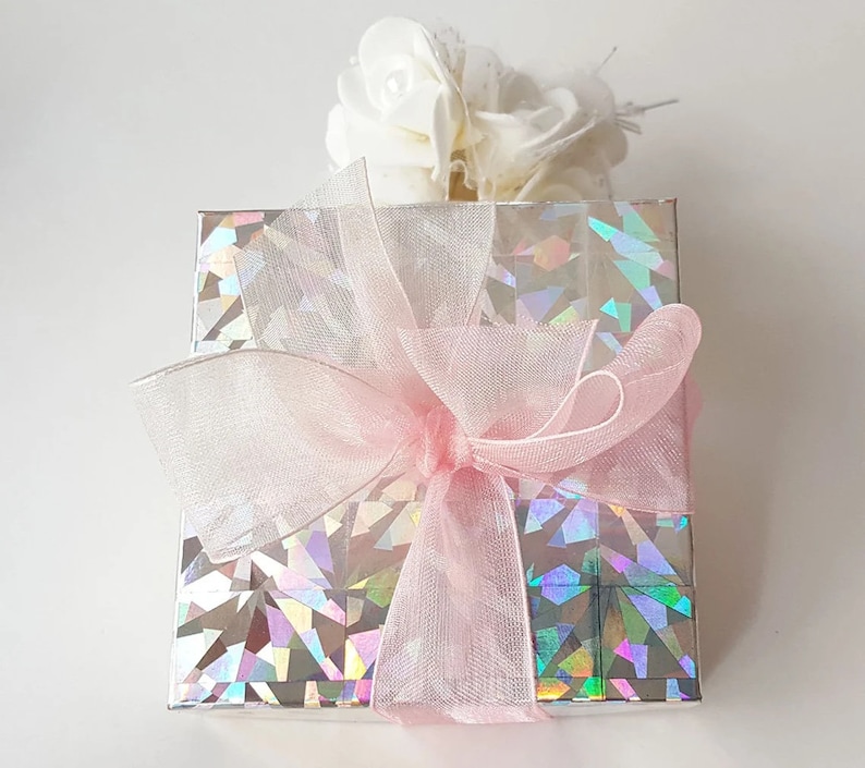 K&ouml;nnte beinhalten: Eine Geschenkbox, verpackt in schimmerndes silbernes Papier mit einer rosa Schleife aus Satin. Die Box ist mit einem wei&szlig;en Blumenarrangement verziert.