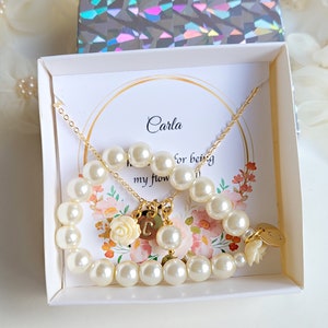 K&ouml;nnte beinhalten: Eine wei&szlig;e Geschenkbox mit einem goldenen Perlen-Halsketten- und Armband-Set. Die Box enth&auml;lt eine Karte mit der Aufschrift "Carla, daf&uuml;r, dass du meine Blume bist".