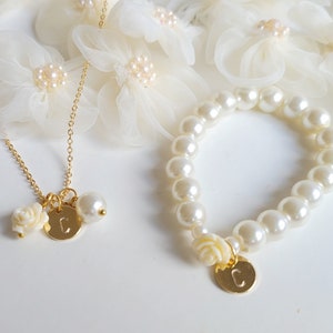 K&ouml;nnte beinhalten: Ein goldenes Halsketten- und Armband-Set mit Perlenakzenten und einem Anh&auml;nger mit dem Buchstaben "C". Die Anh&auml;nger sind eine kleine wei&szlig;e Blume und eine runde Scheibe mit dem Buchstaben "C".